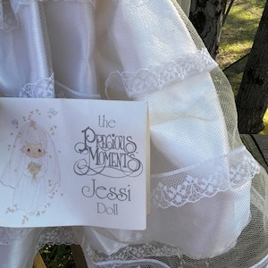 Może przedstawiać: Biała lalka Jessi Precious Moments, z białą sukienką i welonem. Widoczne jest opakowanie lalki, z ilustracją panny młodej i napisem "Precious Moments Jessi Doll."