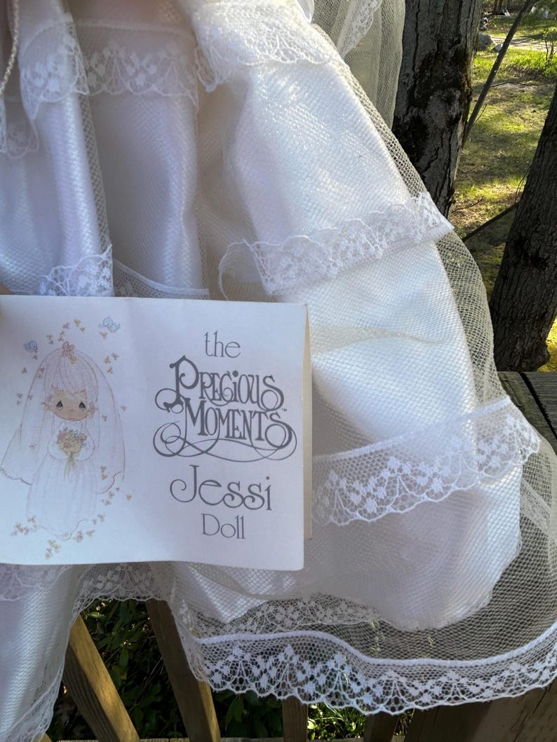Może przedstawiać: Biała lalka Jessi Precious Moments, z białą sukienką i welonem. Widoczne jest opakowanie lalki, z ilustracją panny młodej i napisem "Precious Moments Jessi Doll."