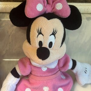 May include: A Minnie Mouse soft toy. The toy has a pink dress with white polka dots, a matching bow with white polka dots, and black ears. The doll has white gloves and a smiling expression.
