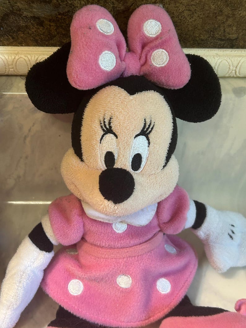 May include: A Minnie Mouse soft toy. The toy has a pink dress with white polka dots, a matching bow with white polka dots, and black ears. The doll has white gloves and a smiling expression.