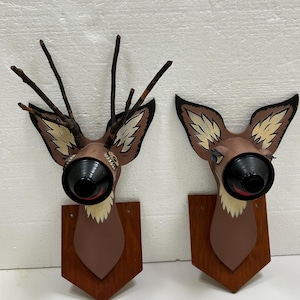 May include: Two brown deer head wall decorations made from recycled materials. The deer have painted eyes, noses, and antlers. The deer are mounted on wooden plaques.