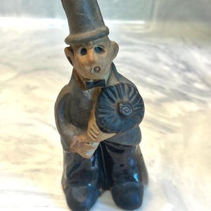 May include: A ceramic figurine of a man in a top hat and suit, holding a dark-colored object. The figure has a neutral skin tone, a black bow tie, and black shoes. The figurine is a decorative item.