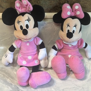 May include: Two Minnie Mouse plush toys. Each toy features black ears, a pink bow with white spots, and a pink dress with white polka dots. The toys have white gloves and pink shoes.