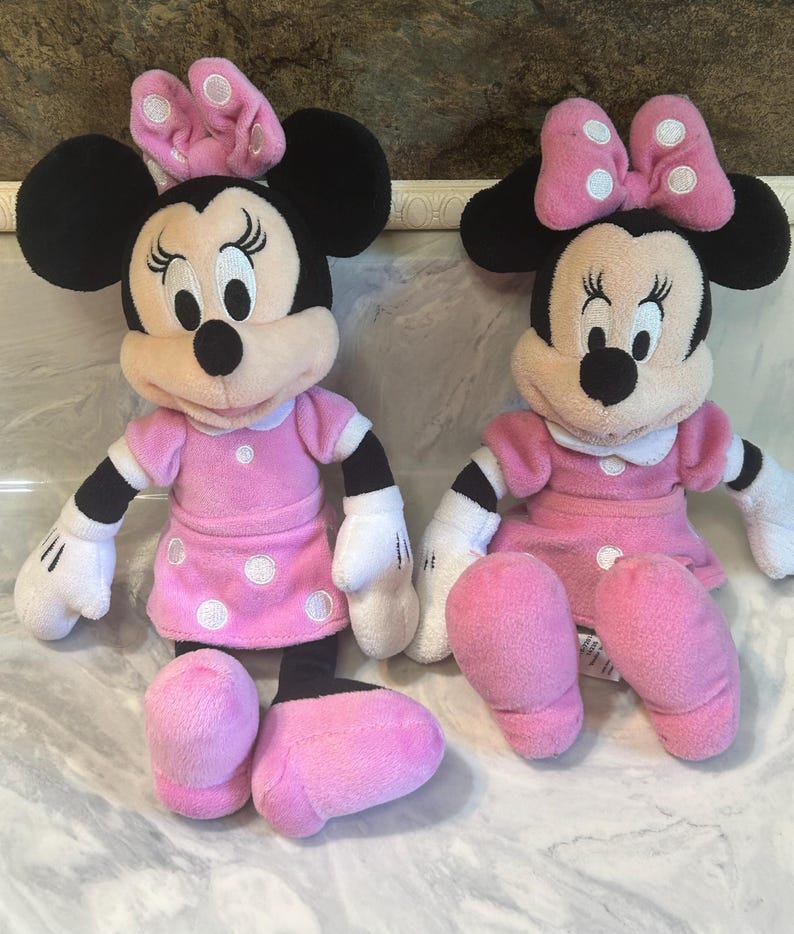 May include: Two Minnie Mouse plush toys. Each toy features black ears, a pink bow with white spots, and a pink dress with white polka dots. The toys have white gloves and pink shoes.