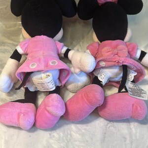 May include: Two Minnie Mouse soft toys. Each toy has black ears, a pink bow, and a pink dress. The toys have white gloves, pink shoes, and black and white striped legs. The toys are facing away from the camera.