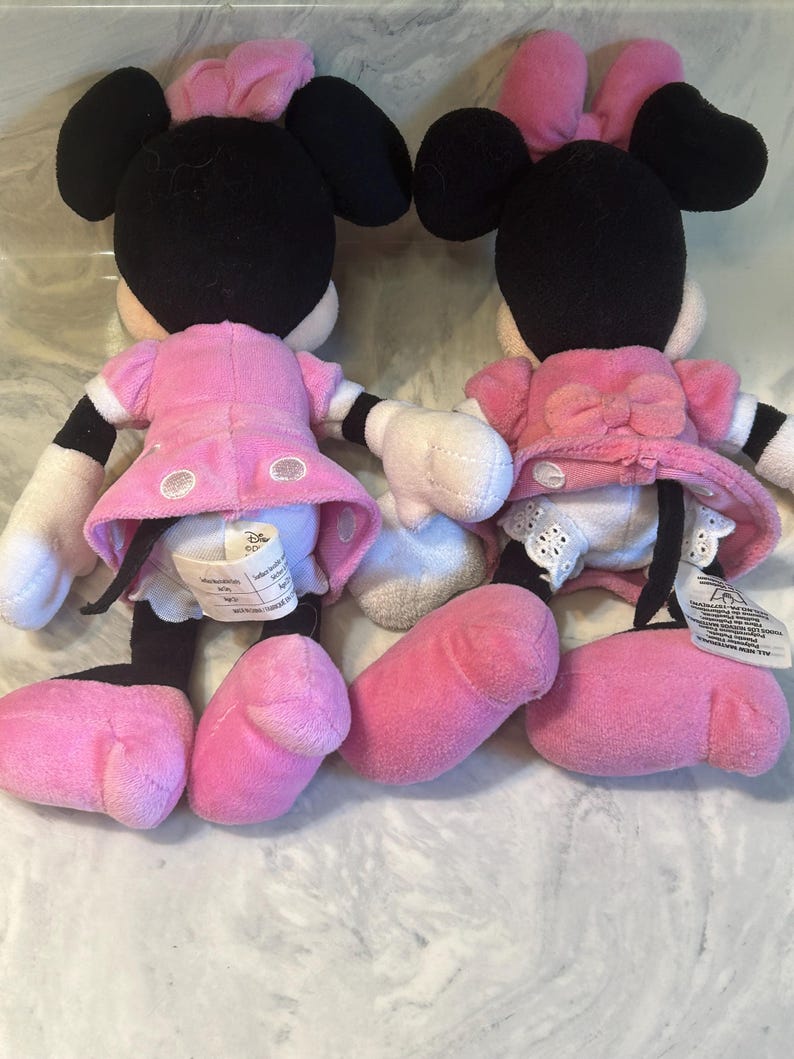 May include: Two Minnie Mouse soft toys. Each toy has black ears, a pink bow, and a pink dress. The toys have white gloves, pink shoes, and black and white striped legs. The toys are facing away from the camera.