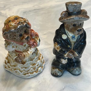 May include: Two ceramic bear figurines, a bride and groom. The bride bear wears a white dress with gold trim and holds a bouquet. The groom bear is in a black suit and top hat. Both have detailed facial features.