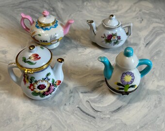 May include: Four miniature porcelain teapots with floral designs. The teapots are in various colors, including pink, blue, and white. The teapots have gold accents and are decorated with flowers and other details.