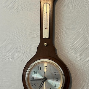 May include: A dark brown wooden wall clock with a thermometer and barometer. The clock face is silver with black numerals and hands. The thermometer displays temperature readings in Fahrenheit. The clock is mounted on a light gray wall.