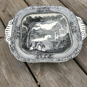 May include: A black and white ceramic serving dish with a raised rim and ornate design. The dish features a landscape scene with a castle, trees, and figures. The dish has two handles on the sides.