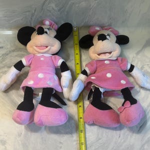 May include: Two plush Minnie Mouse dolls. Each doll has a pink dress with white polka dots, black ears, and a pink bow. The dolls have white gloves and pink shoes. A yellow ruler is placed between the dolls.