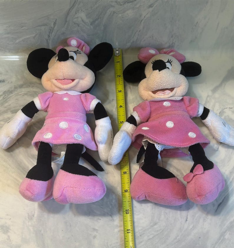 May include: Two plush Minnie Mouse dolls. Each doll has a pink dress with white polka dots, black ears, and a pink bow. The dolls have white gloves and pink shoes. A yellow ruler is placed between the dolls.