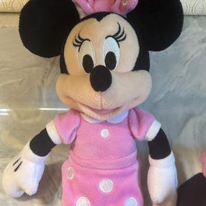 May include: A Minnie Mouse soft toy. The toy has black ears, a pink bow with white polka dots, and a pink dress with white polka dots. It has white gloves and black arms. The background is a light colour.
