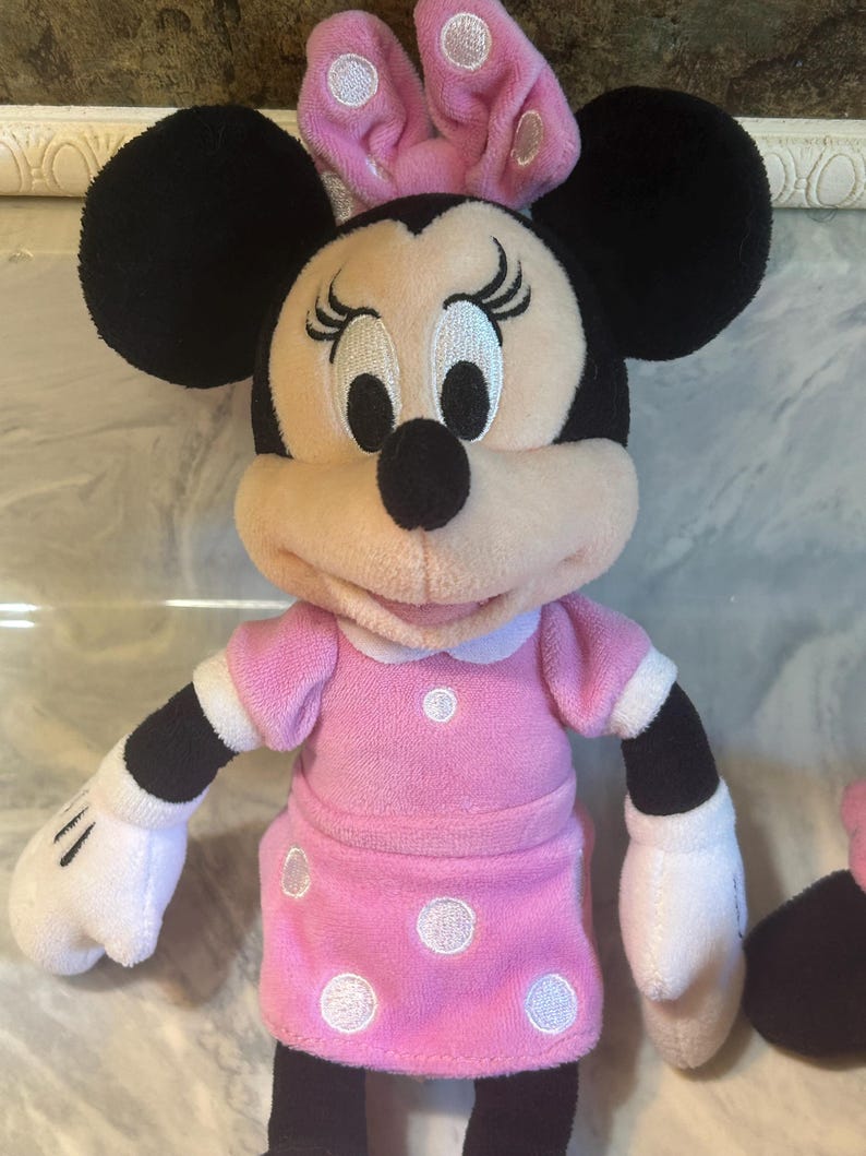 May include: A Minnie Mouse soft toy. The toy has black ears, a pink bow with white polka dots, and a pink dress with white polka dots. It has white gloves and black arms. The background is a light colour.