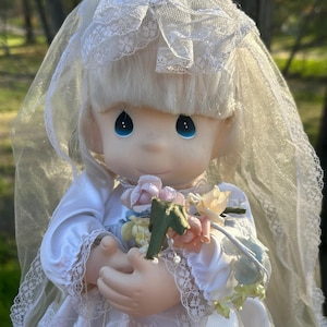 Może przedstawiać: Porcelanowa lalka ubrana jako panna młoda, w białej satynowej sukni z koronkowymi wykończeniami i pasującym welonem. Lalka ma niebieskie oczy i trzyma bukiet kwiat&oacute;w w pastelowych kolorach. Włosy lalki są blond.