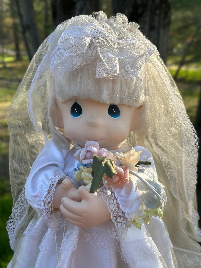 Może przedstawiać: Porcelanowa lalka ubrana jako panna młoda, w białej satynowej sukni z koronkowymi wykończeniami i pasującym welonem. Lalka ma niebieskie oczy i trzyma bukiet kwiat&oacute;w w pastelowych kolorach. Włosy lalki są blond.