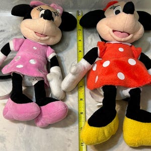May include: Two plush Minnie Mouse dolls. One doll wears a pink dress with white polka dots, a pink bow, and pink shoes. The other doll wears a red dress with white polka dots, a red bow, and yellow shoes. Both dolls have black ears.