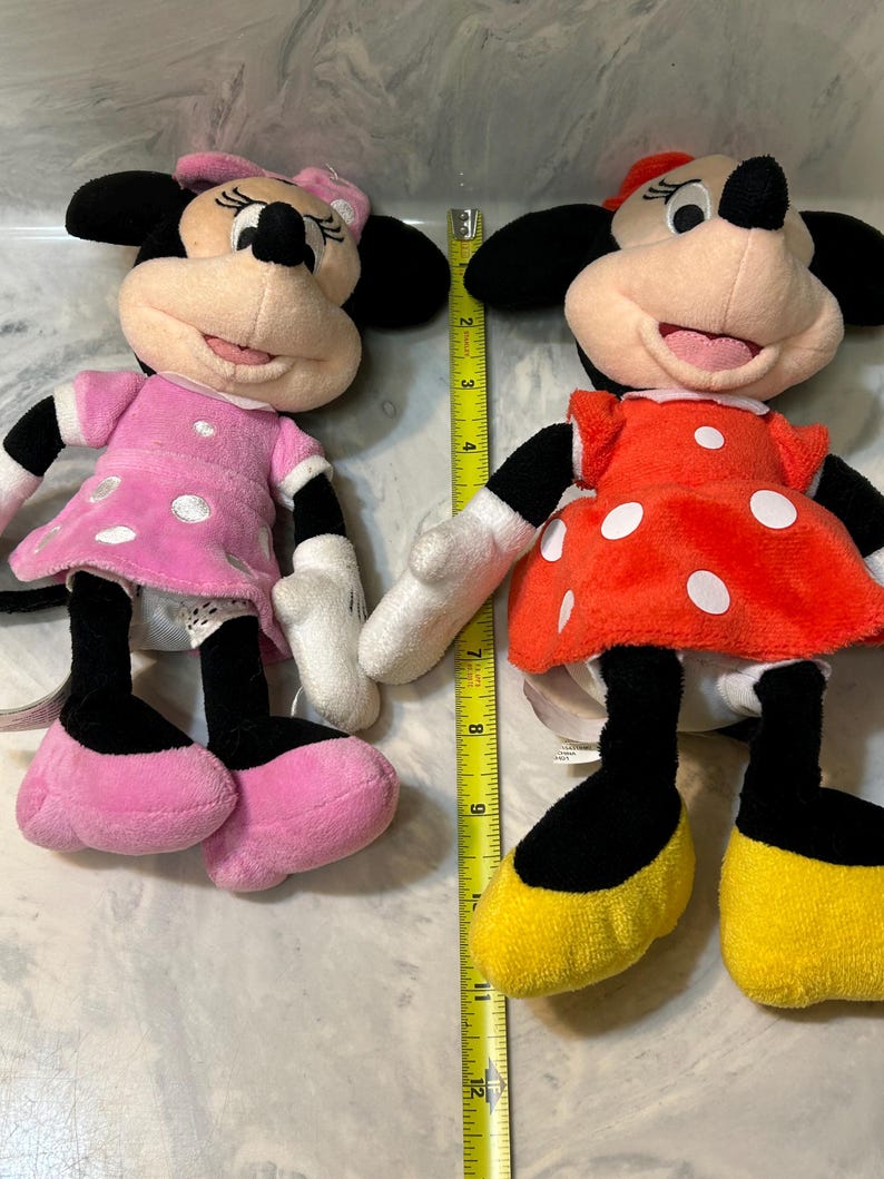 May include: Two plush Minnie Mouse dolls. One doll wears a pink dress with white polka dots, a pink bow, and pink shoes. The other doll wears a red dress with white polka dots, a red bow, and yellow shoes. Both dolls have black ears.