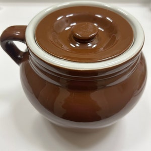 May include: A brown ceramic crock with a lid. The crock has a white rim around the top and a white rim around the lid. The lid has a small knob on top.