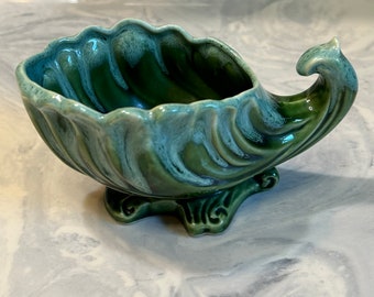 May include: A ceramic, shell-shaped decorative bowl. The bowl is a deep green color with a lighter, blue-green glaze on the edges. It has a curved, wave-like design and sits on three small feet. The bowl's design is reminiscent of ocean waves.