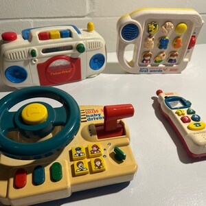 May include: Four Fisher-Price toys for babies and toddlers. A white and red toy with a steering wheel, a white and blue toy with buttons and a red lever, a white and red toy phone with buttons, and a white and yellow toy with a handle and buttons that says "First Words".