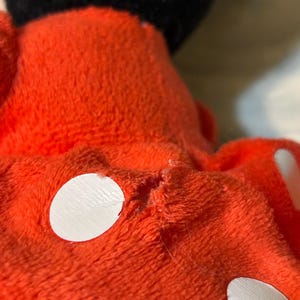 May include: Close-up of a plush toy's red fabric with white circular accents. The toy's black head is visible in the background. The fabric appears worn, with visible stitching and minor damage.