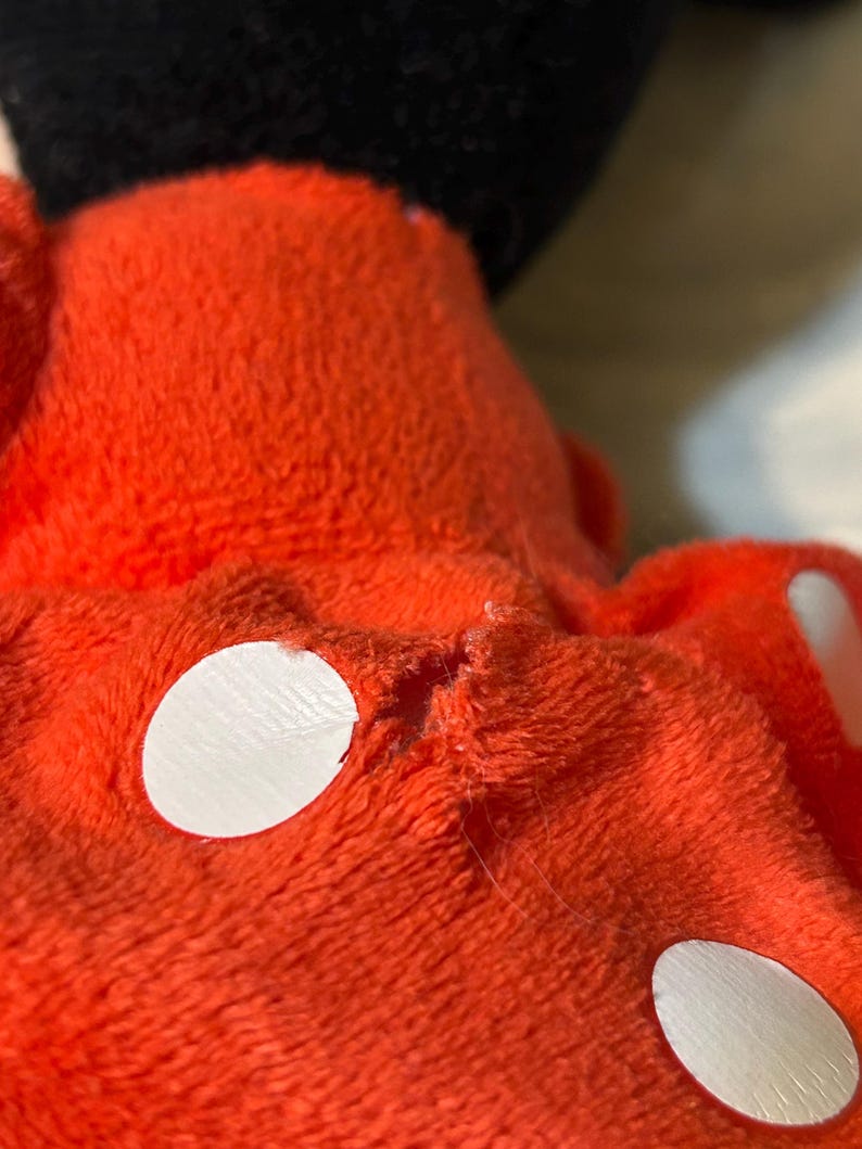 May include: Close-up of a plush toy's red fabric with white circular accents. The toy's black head is visible in the background. The fabric appears worn, with visible stitching and minor damage.