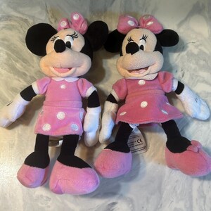 May include: Two Minnie Mouse plush dolls. Each doll has black ears, a pink bow, and a pink dress with white polka dots. The dolls have black legs, pink shoes, and white gloves. The dolls are standing side by side.