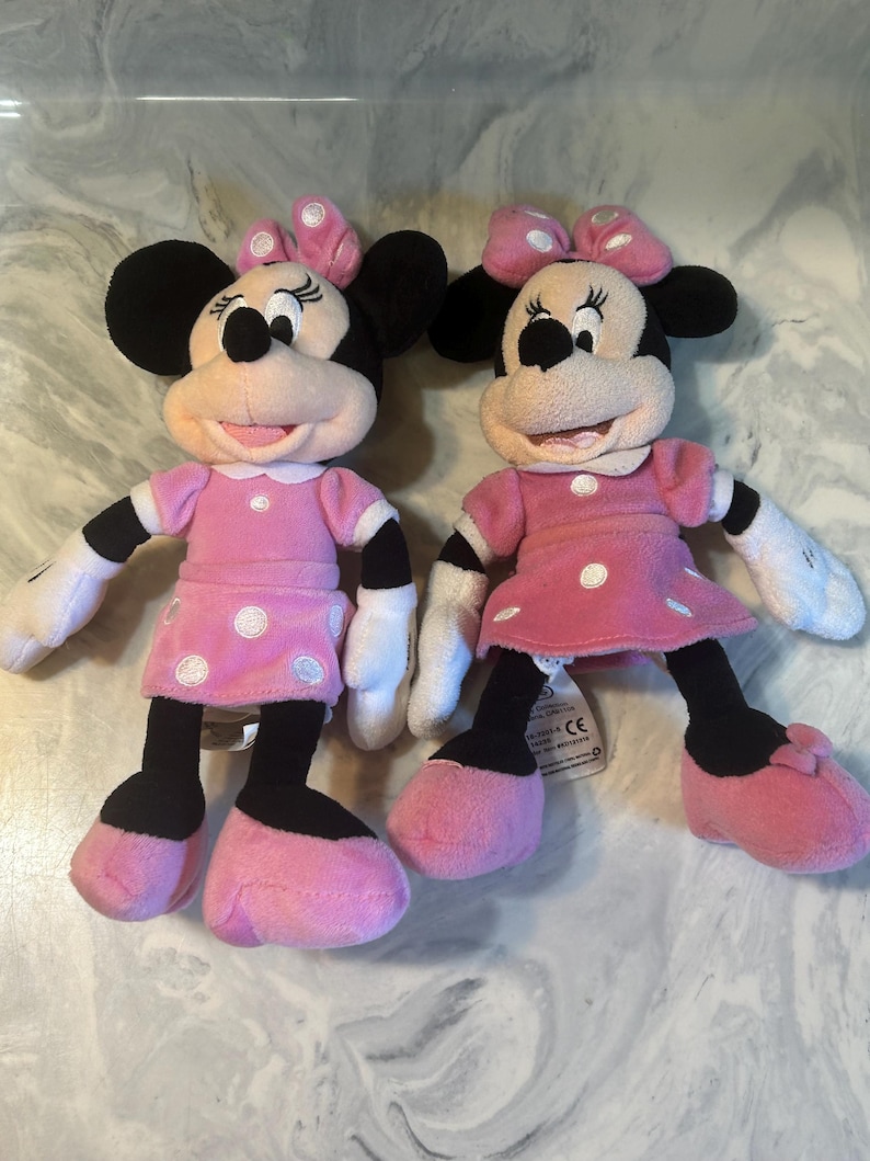 May include: Two Minnie Mouse plush dolls. Each doll has black ears, a pink bow, and a pink dress with white polka dots. The dolls have black legs, pink shoes, and white gloves. The dolls are standing side by side.