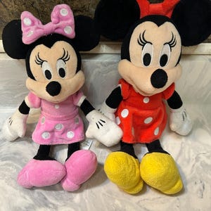 May include: Two plush Minnie Mouse dolls. One wears a pink dress with white polka dots and a matching bow. The other doll is dressed in a red dress with white polka dots and a red bow. Both have black ears, black eyes, and yellow shoes.