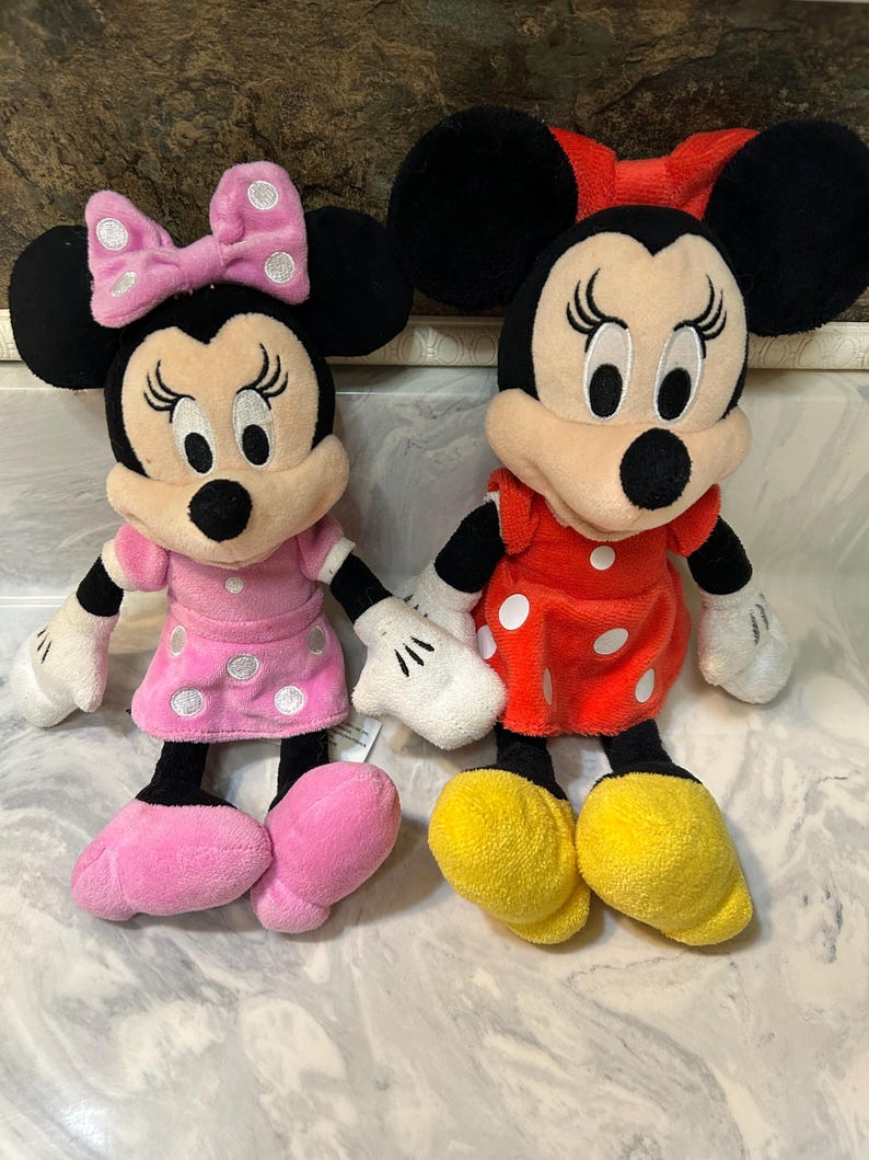 May include: Two plush Minnie Mouse dolls. One wears a pink dress with white polka dots and a matching bow. The other doll is dressed in a red dress with white polka dots and a red bow. Both have black ears, black eyes, and yellow shoes.
