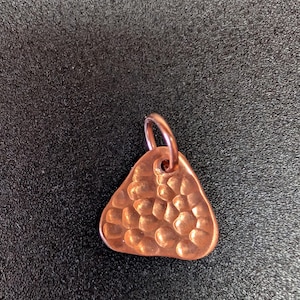 May include: A copper-colored metal pendant with a triangular shape and rounded corners. The surface features a hammered, dimpled texture throughout. A small, circular metal jump ring is attached to the top of the pendant for hanging purposes.