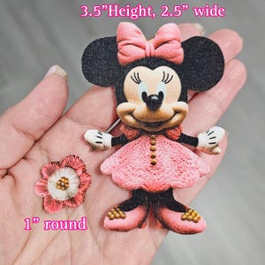 May include: A pink and black fabric applique of a cartoon mouse wearing a pink dress and a pink bow. The applique is 3.5 inches tall and 2.5 inches wide. A pink fabric flower applique is 1 inch in diameter.