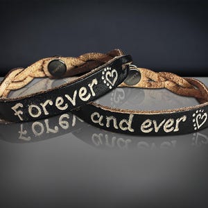 Puede incluir: Dos pulseras de cuero negro con las palabras "Forever and ever" en letras blancas. Las pulseras tienen una banda trenzada de cuero marr&oacute;n claro. Cada pulsera tiene un s&iacute;mbolo de coraz&oacute;n.