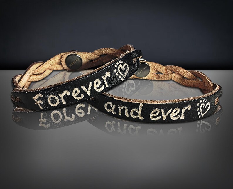 Puede incluir: Dos pulseras de cuero negro con las palabras "Forever and ever" en letras blancas. Las pulseras tienen una banda trenzada de cuero marr&oacute;n claro. Cada pulsera tiene un s&iacute;mbolo de coraz&oacute;n.