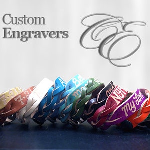 Puede incluir: Una colecci&oacute;n de pulseras de cuero trenzado en varios colores, cada una con texto grabado. Las pulseras se exhiben sobre una superficie oscura contra un fondo claro. El texto "Custom Engravers" es visible.