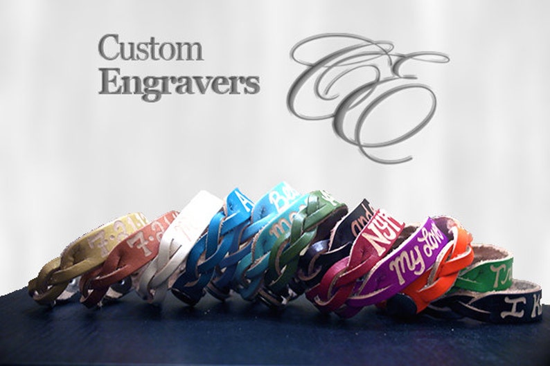 Puede incluir: Una colecci&oacute;n de pulseras de cuero trenzado en varios colores, cada una con texto grabado. Las pulseras se exhiben sobre una superficie oscura contra un fondo claro. El texto "Custom Engravers" es visible.