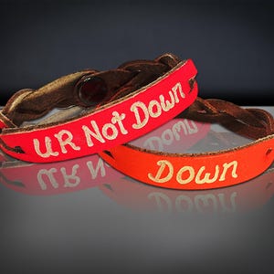 Puede incluir: Dos pulseras de cuero. Una es roja con las palabras "UR Not Down" en letras doradas. La otra es naranja con la palabra "Down" en letras doradas. Ambas pulseras tienen una banda de cuero trenzado marr&oacute;n.