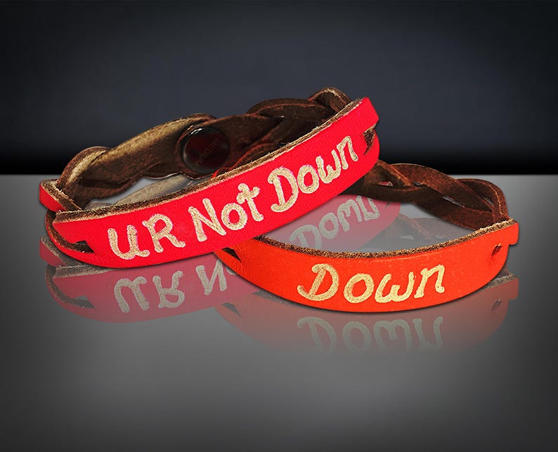 Puede incluir: Dos pulseras de cuero. Una es roja con las palabras "UR Not Down" en letras doradas. La otra es naranja con la palabra "Down" en letras doradas. Ambas pulseras tienen una banda de cuero trenzado marr&oacute;n.
