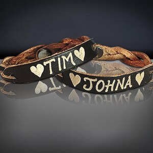 Puede incluir: Dos pulseras de cuero marr&oacute;n oscuro con los nombres "TIM" y "JOHNA" en letras doradas, cada una con un s&iacute;mbolo de coraz&oacute;n dorado. Las pulseras tienen una banda de cuero trenzado.
