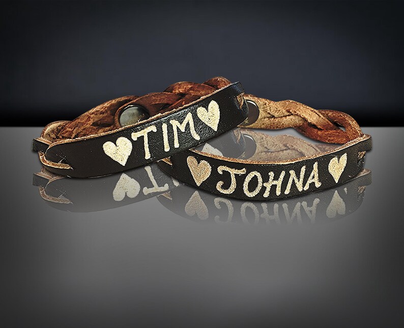Puede incluir: Dos pulseras de cuero marr&oacute;n oscuro con los nombres "TIM" y "JOHNA" en letras doradas, cada una con un s&iacute;mbolo de coraz&oacute;n dorado. Las pulseras tienen una banda de cuero trenzado.