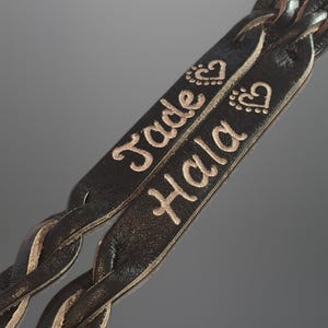 Puede incluir: Primer plano de dos correas de cuero negro trenzado. Las correas est&aacute;n grabadas con los nombres "Jade" y "Hala" en color marr&oacute;n claro, junto con dise&ntilde;os de corazones y puntos. El cuero tiene una apariencia texturizada.