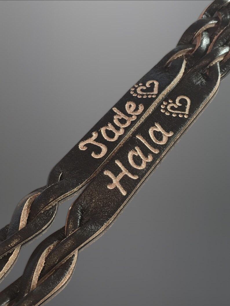 Puede incluir: Primer plano de dos correas de cuero negro trenzado. Las correas est&aacute;n grabadas con los nombres "Jade" y "Hala" en color marr&oacute;n claro, junto con dise&ntilde;os de corazones y puntos. El cuero tiene una apariencia texturizada.