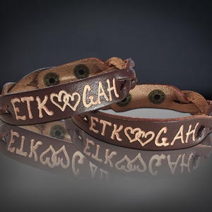 Puede incluir: Dos pulseras de cuero marr&oacute;n con las letras grabadas "ETK" y "GAH" separadas por dos corazones. Las pulseras tienen un dise&ntilde;o trenzado y cierres de metal. El fondo es oscuro.