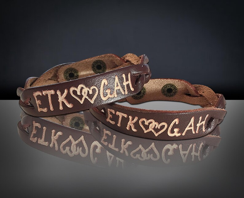 Puede incluir: Dos pulseras de cuero marr&oacute;n con las letras grabadas "ETK" y "GAH" separadas por dos corazones. Las pulseras tienen un dise&ntilde;o trenzado y cierres de metal. El fondo es oscuro.