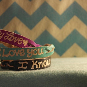 Puede incluir: Tres pulseras de cuero apiladas. La pulsera superior es morada y dice "my Love" con un coraz&oacute;n. La pulsera del medio es turquesa y dice "I Love You". La pulsera inferior es negra y dice "I Know".