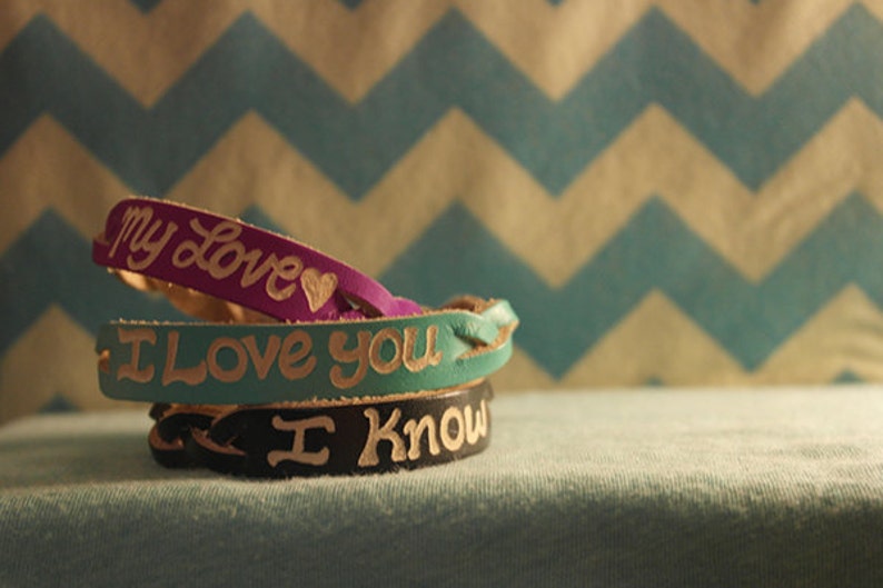 Puede incluir: Tres pulseras de cuero apiladas. La pulsera superior es morada y dice "my Love" con un coraz&oacute;n. La pulsera del medio es turquesa y dice "I Love You". La pulsera inferior es negra y dice "I Know".