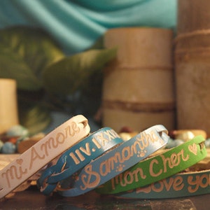 Puede incluir: Una colecci&oacute;n de pulseras de cuero personalizadas en varios colores, incluyendo crema, azul y verde. Cada pulsera presenta texto grabado, con frases como "Mi Amore" y nombres. Las pulseras se exhiben con elementos decorativos de bamb&uacute;.