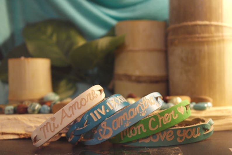 Puede incluir: Una colecci&oacute;n de pulseras de cuero personalizadas en varios colores, incluyendo crema, azul y verde. Cada pulsera presenta texto grabado, con frases como "Mi Amore" y nombres. Las pulseras se exhiben con elementos decorativos de bamb&uacute;.