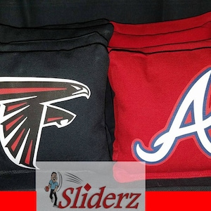 May include: Two cornhole bags, one black with a white and red Atlanta Falcons logo, and one red with a white and blue Atlanta Braves logo. The bags are made of a durable fabric and are designed for outdoor games.